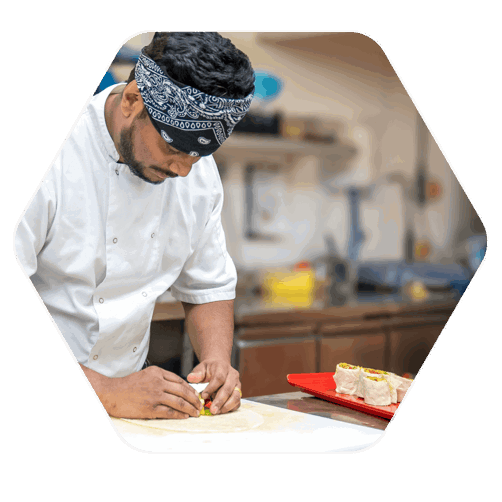 A chef with a black and white bandana on his head. The chef is leaning over this work bench, rolling ingredients into a tortilla wrap dish