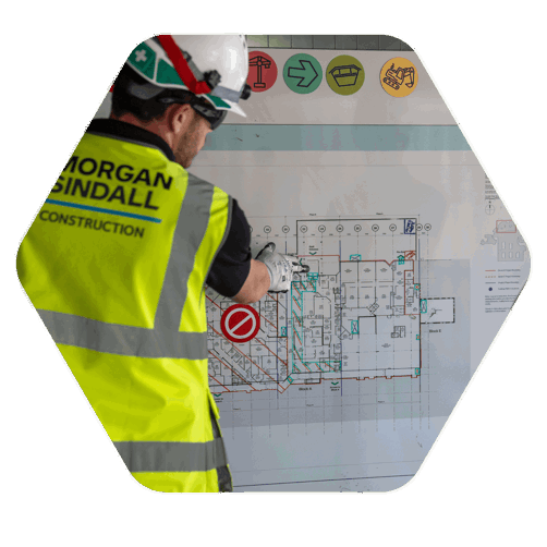 A construction worker with a high vis jacket and hard hat on. His back is turned and he is pointing to a structure that is on a blue print hanging in front of him on a board
