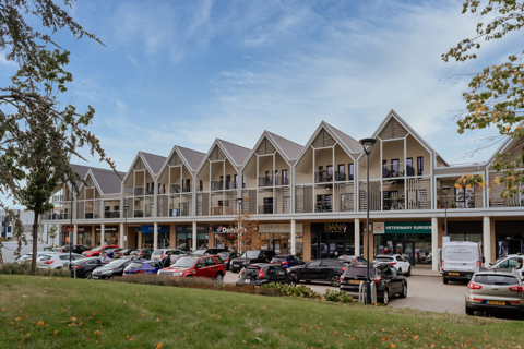 A row of new build flats with shops underneath on the ground floor, cars park in front of the structure