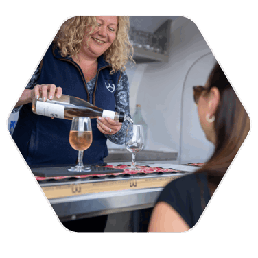 A blonde haired woman smiling in a mobile bar vehicle pouring a pink coloured wine from a bottle into a glass which is positioned next to a full glass
